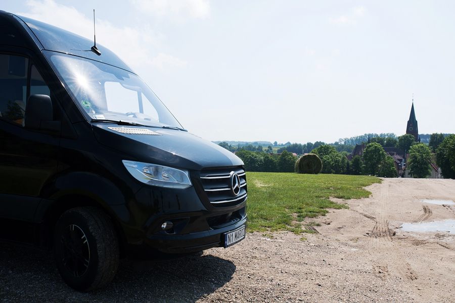 Czarny Mercedes Sprinter VIP do przewozu osób — zdjęcie samochodu zrobione w Gietrzwałdzie, w tle wieża sanktuarium